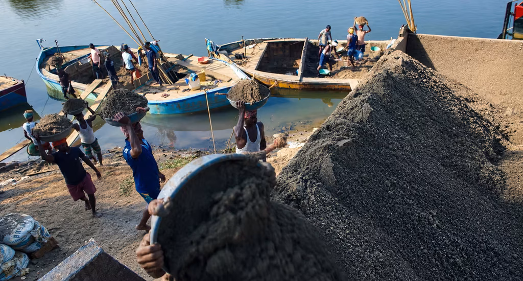 Reporting Live scene featuring laborers carrying sand in baskets beside wooden boats along a riverside.